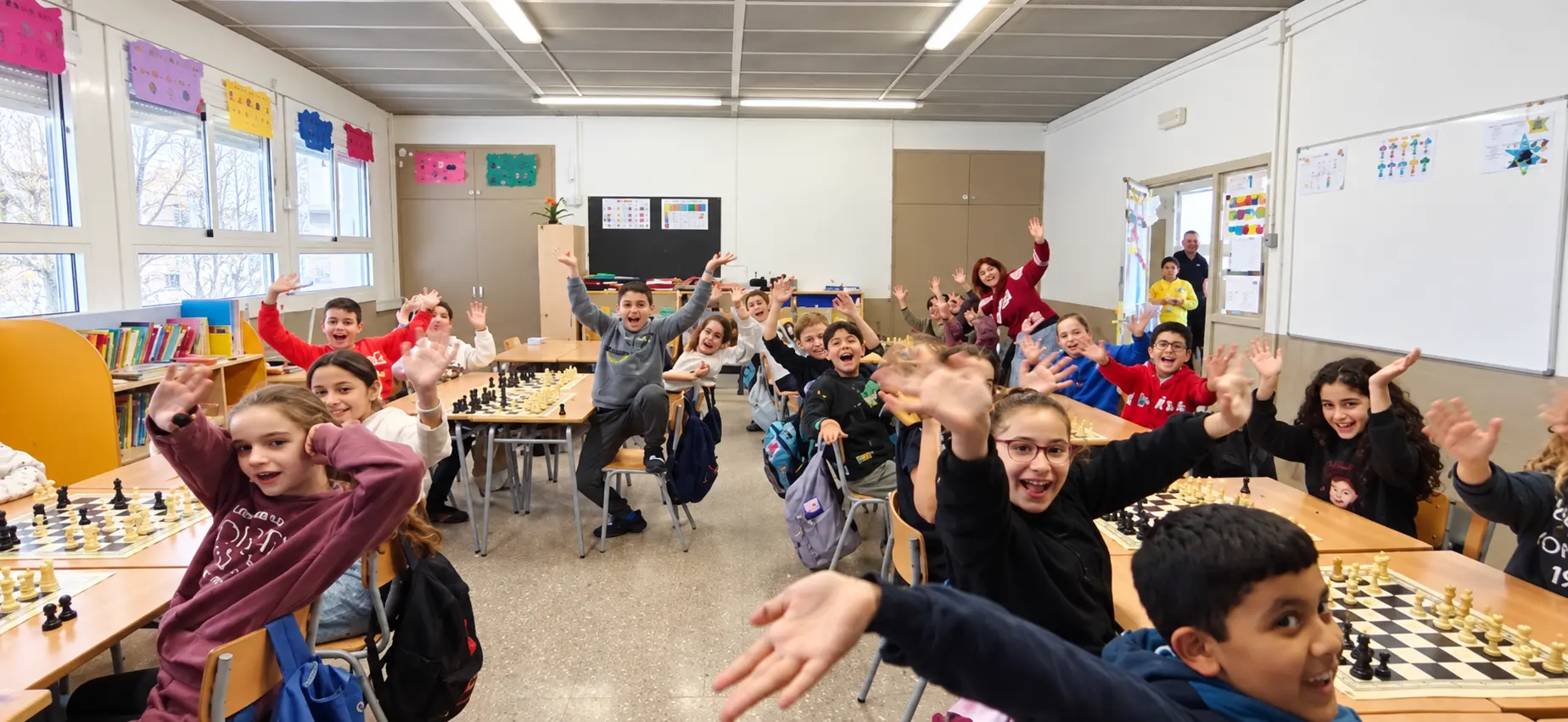 Alumnos saludando a cámara durante un lipdub escolar en un aula con mesas de ajedrez
