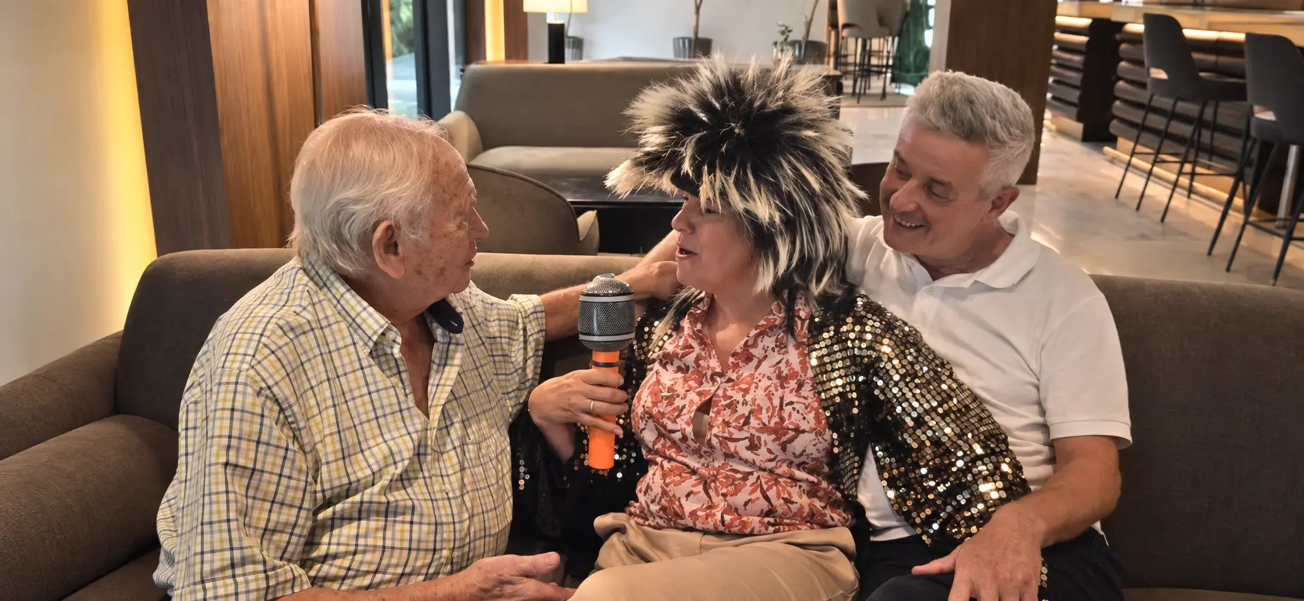Mujer con peluca y micrófono junto a dos invitados durante un videoclip de cumpleaños