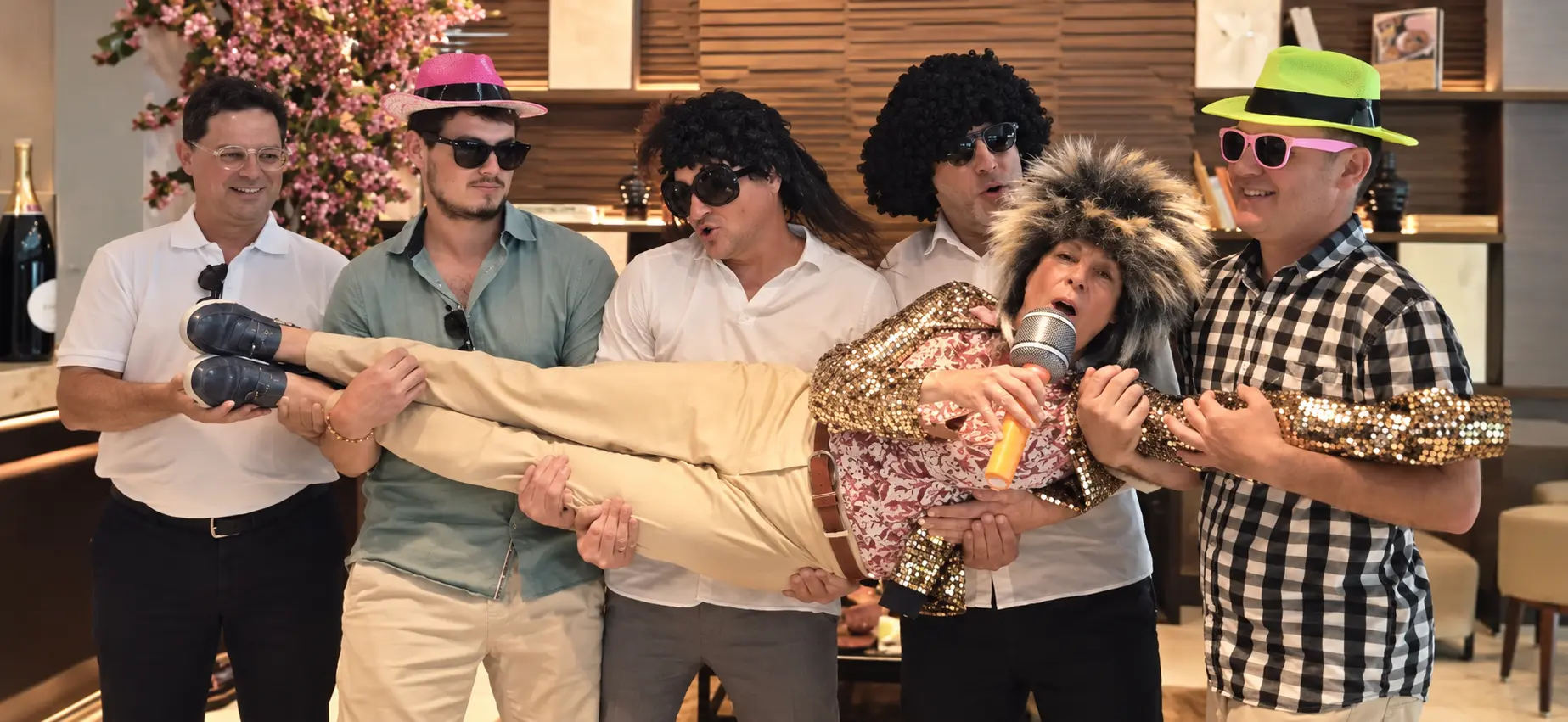 Cinco invitados sosteniendo a una participante con peluca y micrófono en una escena divertida de cumpleaños
